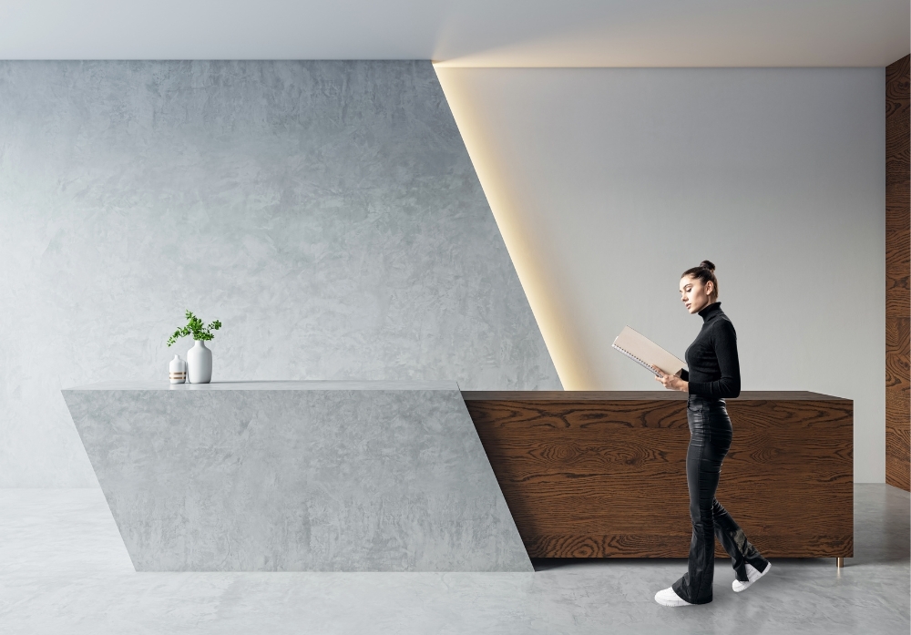 A modern, minimalist reception area featuring a geometric concrete and wood desk. A woman dressed in black, wearing a turtleneck and flared pants, walks past while reading a notebook